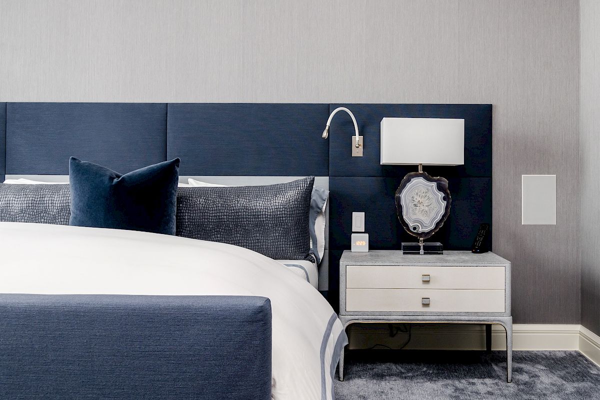 A modern bedroom with a navy headboard, white bedding, a nightstand with a lamp, and decorative pillows is shown.