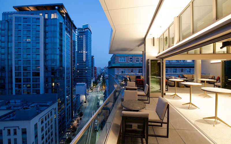 An outdoor balcony with tables and chairs overlooks a cityscape with modern high-rise buildings, capturing the scene during twilight.