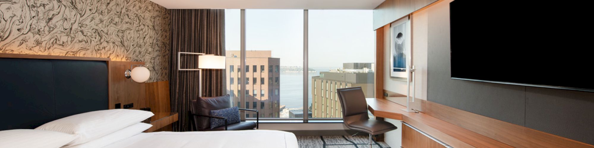 The image shows a modern hotel room with a large bed, desk, chair, flat-screen TV, and a window offering a view of buildings and water.